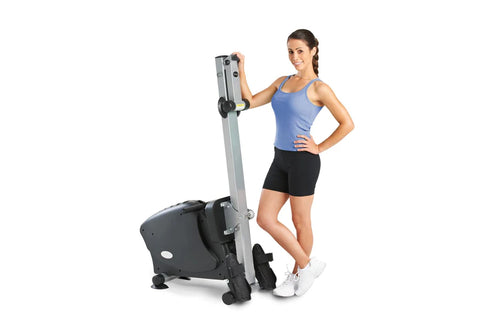 Black and gray stationary rowing machine with extended handle, displayed with woman in blue tank top and black shorts standin