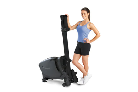 Black and gray stationary rowing machine with extended handle, displayed with woman in blue tank top and black shorts standin