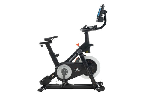 Black NordicTrack S15i indoor exercise bike with attached touchscreen display showing workout content, viewed from side on wh