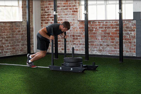 Man pushing black sled on bright green synthetic turf in industrial gym space with brick walls