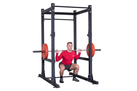 Black power rack with perforated uprights, red weight plates on loaded barbell, man in red shirt performing squat exercise