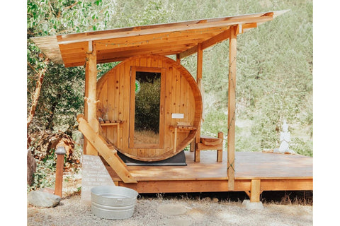 Barrel sauna made of light wood with horizontal planks, open wooden door visible, black metal chimney pipe extending from top