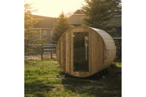 Wooden barrel sauna with rounded cylindrical design and dark glass door, positioned in backyard setting with residential buil