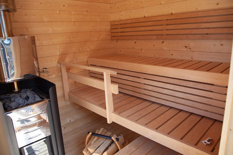 Modern wooden sauna cabin with black roof, glass door, louvered side panels, and green deck foundation on white background