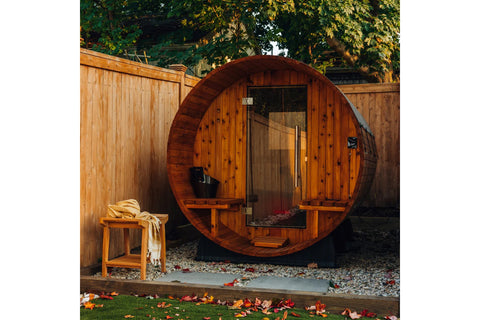 Round barrel sauna with light wood construction, glass door, wooden benches, and bucket visible inside, on black support base