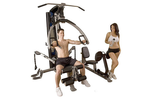 Multi-station home gym with gray and blue frame, featuring chest press, leg press, and cable machine stations, demonstrated b