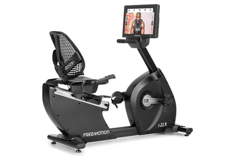 Black Freemotion r22.9 recumbent exercise bike with mesh backrest, foot pedals, and attached digital display screen showing w