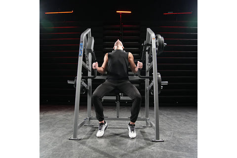 Black plate-loaded multi-station gym equipment with neck exercise attachment, barbell sleeve, and weight plate storage on whi