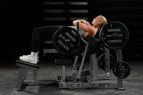 Black plate-loaded glute drive machine with padded seat and backrest, multiple weight plate pegs, and handlebar grips on whit