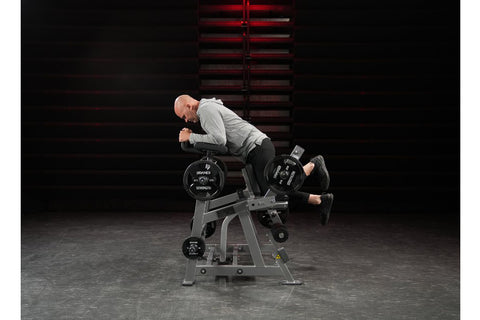 Black plate-loaded leg curl machine with padded seat, backrest, and leg extension arms, multiple weight plate pegs, on white 