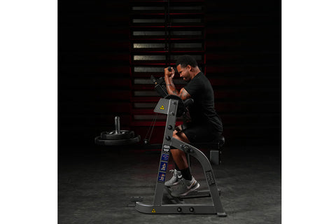Black plate-loaded seated biceps machine with padded seat and arm supports on white background