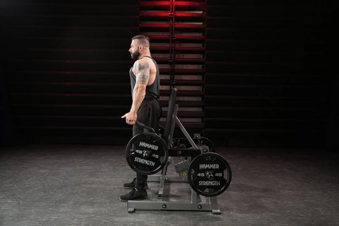 Black plate-loaded shrug machine with padded shoulder support, adjustable arms, and weight plate holders on white background