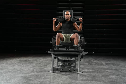 Black plate-loaded V-squat machine with padded seat and backrest, adjustable arm supports, and metal frame on white backgroun