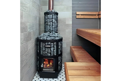Black cylindrical wood-burning sauna stove with gray rocks on top, viewing window showing flames, and Harvia branding on whit