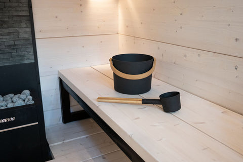 Black steel sauna pail with bamboo handle strap and matching black aluminum ladle with wooden handle, on white background