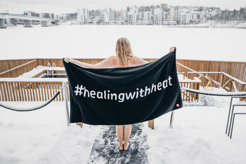 Black sauna towel with white "#healingwithheat" text, held by person at outdoor sauna deck with frozen lake and city skyline
