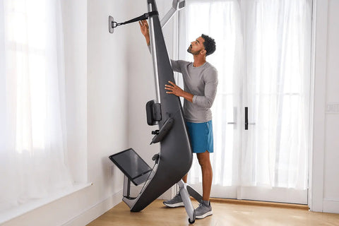 Hydrow Arc Rower with black seat and silver frame, mounted touchscreen displaying water rowing video, on wooden floor in home