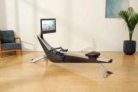 Hydrow Origin rowing machine with black seat and silver frame, featuring mounted tablet display showing workout video, on whi