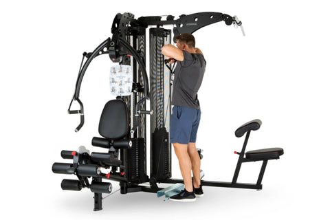 Black multi-station home gym with man demonstrating upper body exercise, featuring adjustable weight stack, leg press platfor