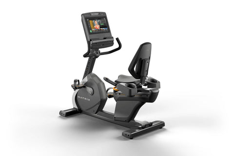 Black and gray Matrix recumbent exercise bike with digital display screen, padded seat, and foot pedals on white background
