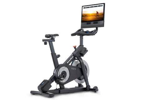 NordicTrack X24 black indoor exercise bike with attached tablet displaying sunset cycling video on white background