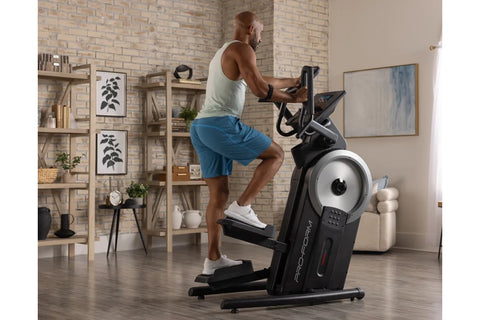 Man in white tank top and blue shorts using black ProForm elliptical machine with silver flywheel in home gym with brick wall