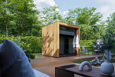 Modern wooden cube sauna with dark interior and windows, situated on composite deck surrounded by landscaping and garden furn