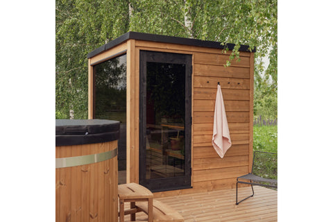 Cedar wood outdoor sauna with black roof and glass door, mounted on wooden deck with ivy-covered wall backdrop