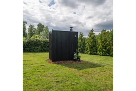 Black vertical wood-slat outdoor sauna with chimney in landscaped garden surrounded by green lawn and evergreen trees