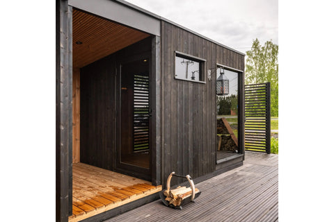 Black vertical wood-slat outdoor sauna with chimney in landscaped garden surrounded by green lawn and evergreen trees