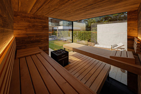 Modern wooden outdoor sauna with vertical slat design positioned on lawn beside contemporary architecture and green hedge wal