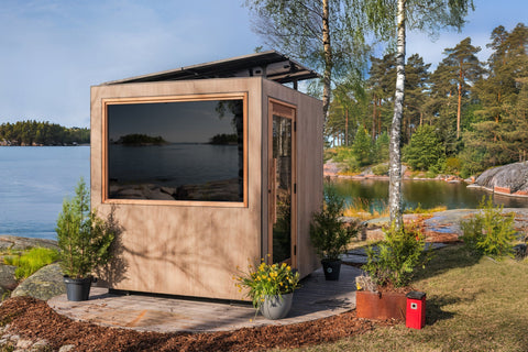 Modern wooden sauna cabin with large dark window, black roof trim, and entry door on lakeside setting surrounded by trees and