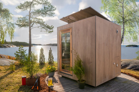 Modern wooden sauna cabin with large dark window, black roof trim, and entry door on lakeside setting surrounded by trees and