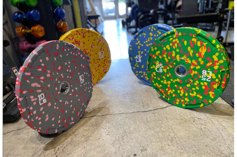 Colorful bumper plates with speckled rubber coating displayed on gym floor, including gray with red flecks, yellow with red f