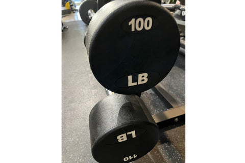 Black fixed dumbbell with white "100 LB" markings and gray textured handle, resting on gym floor
