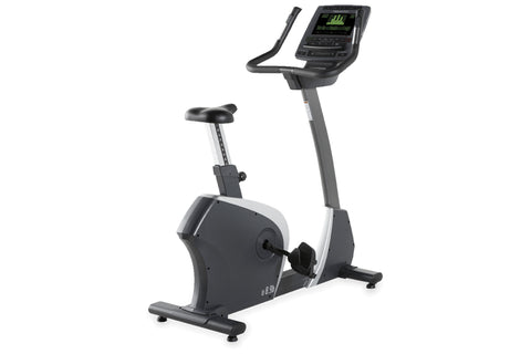 Black and white upright exercise bike with digital display console showing green metrics, foot pedals, and adjustable seat on