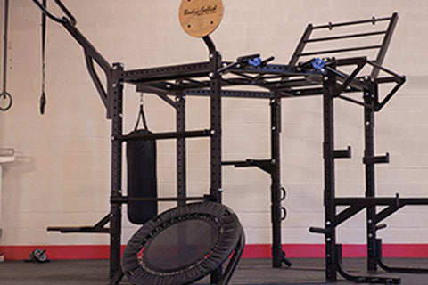 Black multi-function fitness rig with pull-up bar, dip station, and rope attachment, displayed with rebounder trampoline in g