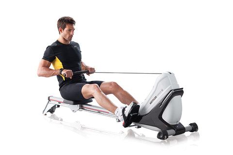 Man using white and black indoor rowing machine with yellow resistance band, wearing black Nike shirt and shorts on white bac