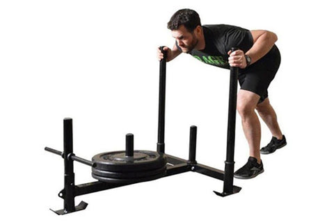 Man using black Warrior Weighted Push Sled with loaded plate, demonstrating forward push motion on white background