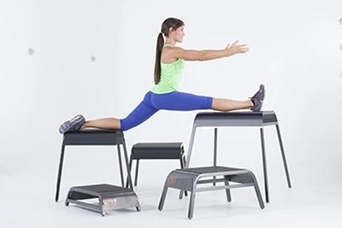 Woman in lime green tank top and blue leggings performing a jump exercise on black and gray plyometric boxes