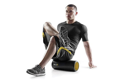 Male model wearing black and yellow athletic shirt and shorts, using black and gold foam roller on leg against white backgrou