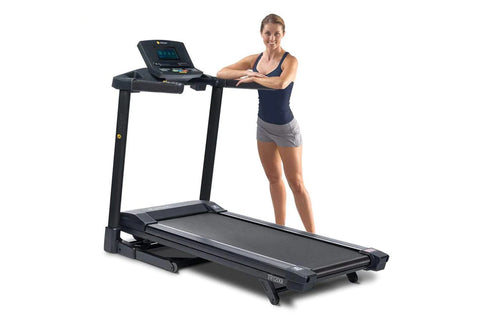 Black folding treadmill with digital display console, shown with woman in navy tank top and gray shorts standing beside it on