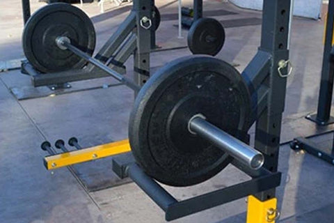 Black rubber weight plate loaded on silver barbell in squat rack with yellow safety tray attachment, indoor gym setting
