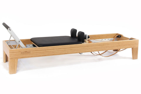 Natural wood pilates reformer with black padded carriage, footbar, and black foot blocks on white background