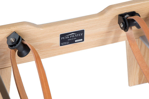 Natural wood pilates reformer with black padded carriage, footbar, and black foot blocks on white background
