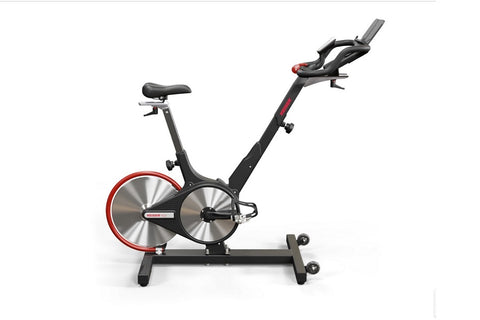Black and red indoor stationary exercise bike with dual flywheels, black handlebars, gray seat post, and wide base on white b