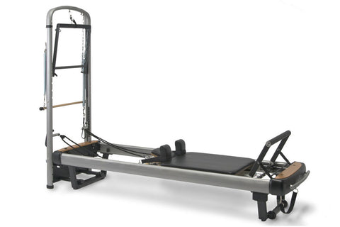 Peak Pilates MVe+ Tower System with gray metal frame, black padded bed, and wooden foot bar on white background