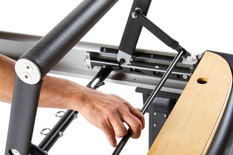 Peak Pilates MVe+ Tower System with gray metal frame, black padded bed, and wooden foot bar on white background