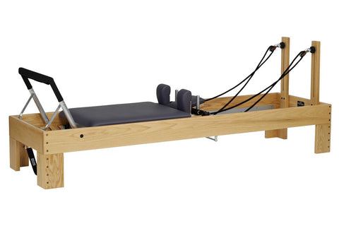 Natural wood Pilates reformer with black padded carriage, footbar, and dual wooden uprights with black rope resistance straps