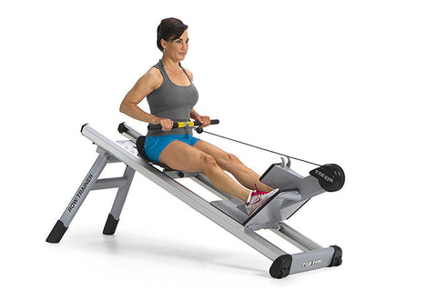 Woman using gray and black Total Gym Row Trainer exercise machine, wearing gray tank top and blue shorts, on white background